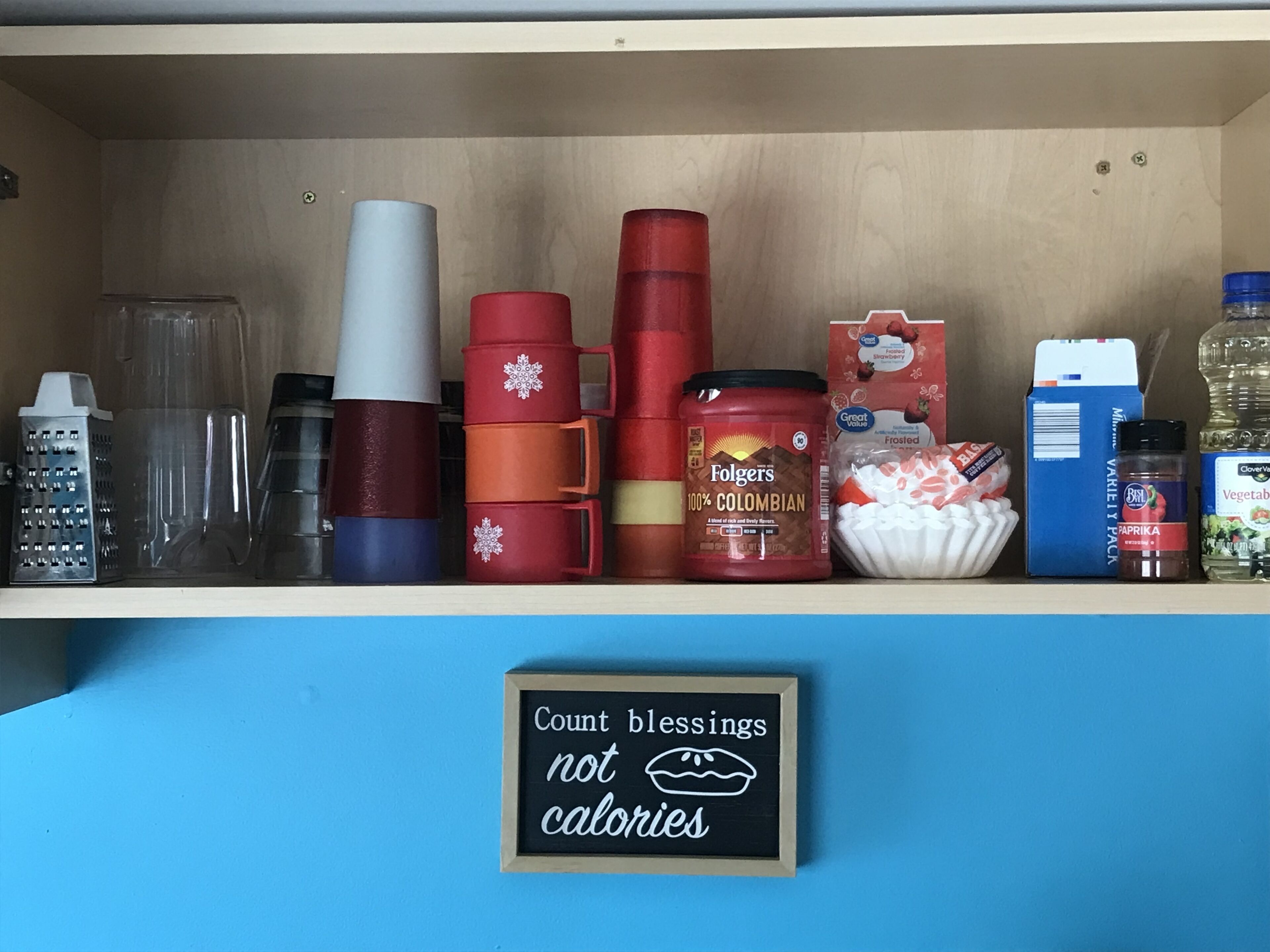 Una estantería con varios utensilios de cocina, como un rallador, tazas, un tarro de café Folgers, un cartón de leche y un letrero que dice 'Cuenta bendiciones, no calorías'.