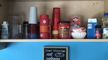 Una estantería con varios utensilios de cocina, como un rallador, tazas, un tarro de café Folgers, un cartón de leche y un letrero que dice 'Cuenta bendiciones, no calorías'.
