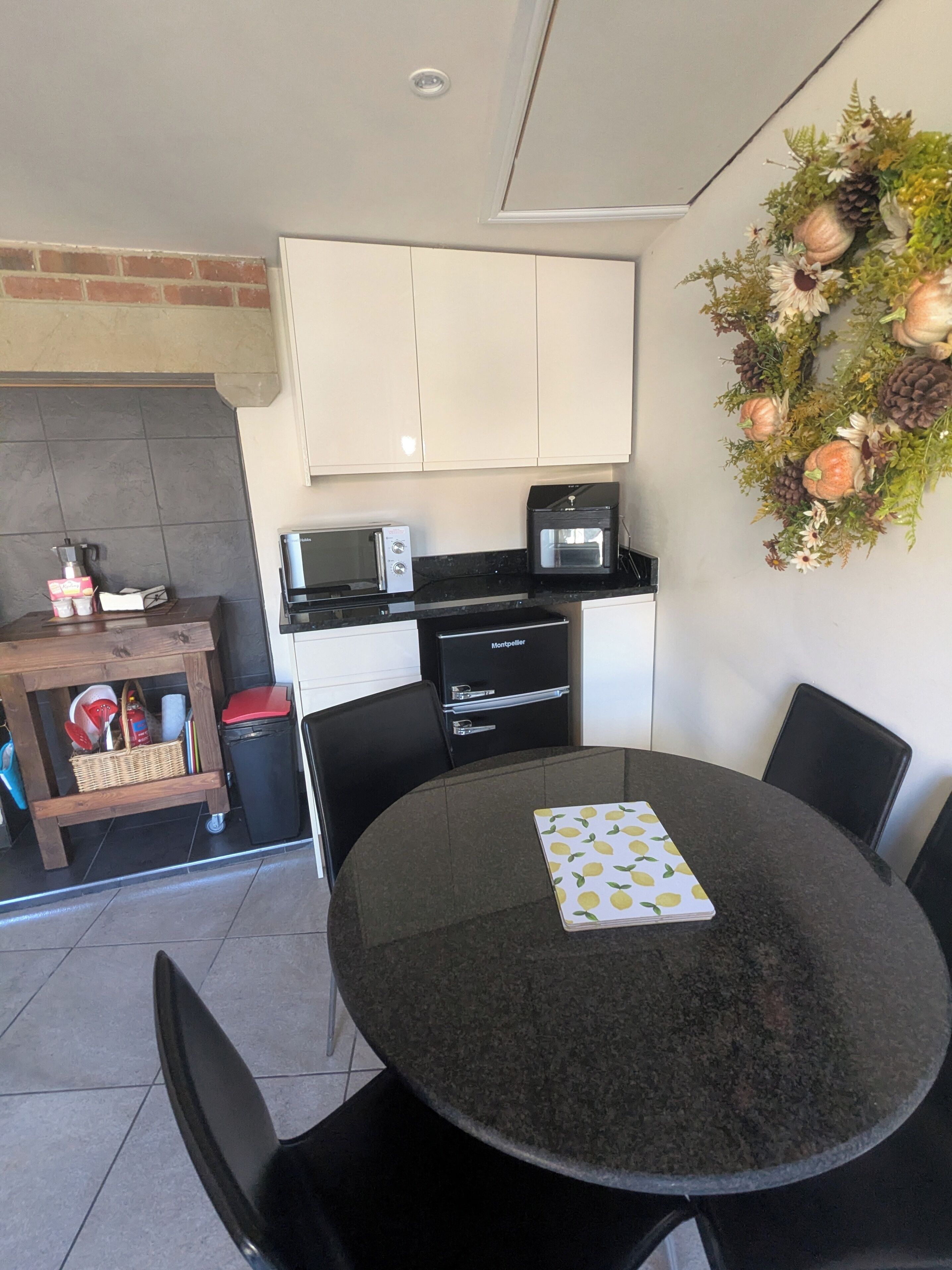 Cocina compacta con armarios blancos, microondas, tostadora y un refrigerador pequeño. En primer plano, una mesa redonda con sillas, y un adorno de pared con piñas y flores.