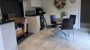 Cocina compacta con microondas, tostadora y refrigerador pequeño. Hay un área de comedor con mesa y sillas, y una corona decorativa en la pared.