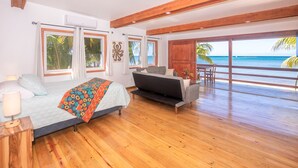 Un dormitorio con una cama, un sofá, suelo de madera y grandes ventanas con vistas a la playa.