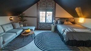 Ein gemütliches Schlafzimmer im Loft-Stil mit schrägverdachter Decke, einem großen Fenster mit gemusterten Vorhängen, einer Couch, einem Bett und einem runden Couchtisch.