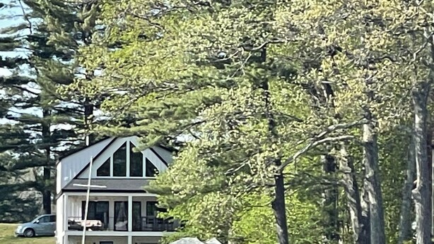 Una casa frente al lago con una edificación principal y una estructura más pequeña, rodeada de árboles y una orilla rocosa.