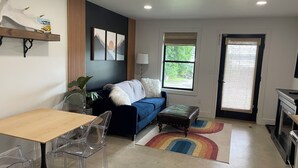 Una sala de estar moderna con un sofá azul, una mesa de comedor de madera y sillas transparentes. Hay una alfombra colorida en el suelo, una ventana con vistas a vegetación y una puerta con cortina.