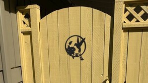 A yellow wooden gate with a circular emblem featuring a bird and a leaf. - Maple Valley Hummingbird Studio (Maple Valley)