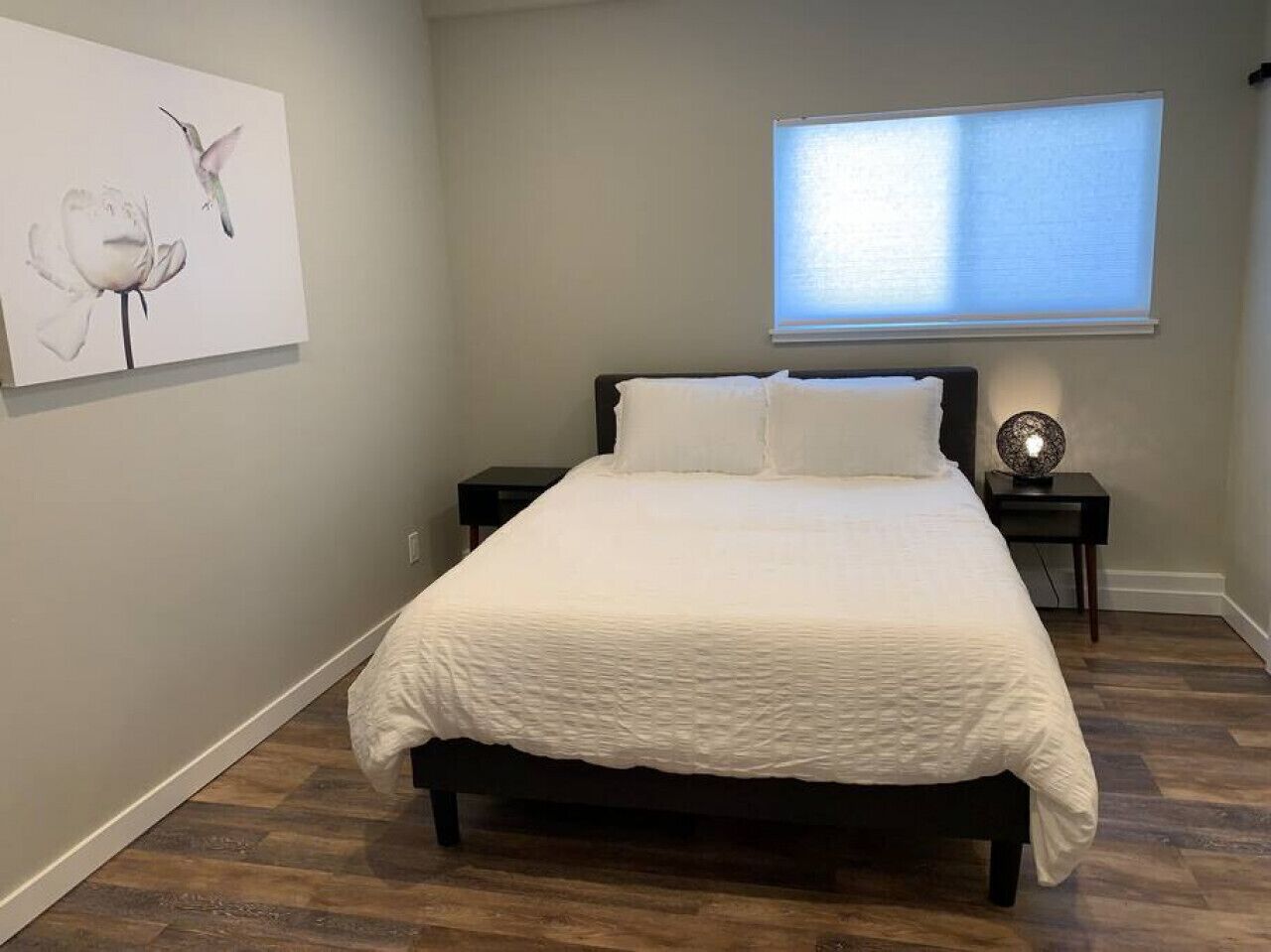 A bedroom with a bed, a nightstand, a wall-mounted lamp, and a window with blinds.