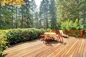 A wooden deck with a table and chairs, surrounded by dense greenery and tall trees. - Central Sammamish Home With Private Setting (Sammamish)