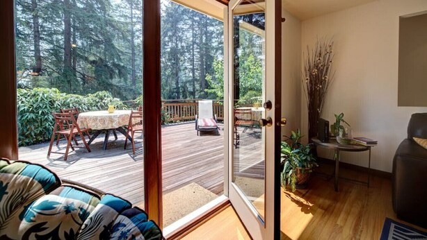 A room with a sliding glass door leading to an outdoor deck with a table and chairs, surrounded by greenery.