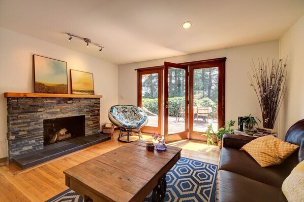 A living room with a stone fireplace, a wooden coffee table, a brown leather sofa, and a large window with a view of greenery. - Central Sammamish Home With Private Setting (Sammamish)