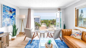A modern living room with a view of the beach, a sofa, chairs, a coffee table, and a painting on the wall. - Gertrude's Siesta | Oceanview, 12-steps Beach, 4-min trolley to Village, tiki-bar, ping-pong! (Siesta Key)
