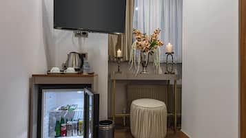 Habitació d'hotel compacta amb televisió de pantalla plana, nevera minibar plena de begudes, taula petita amb cafetera, gerra amb flors i una otomana rodona.