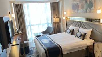 Habitación de hotel con una cama grande, un escritorio con televisor de pantalla plana, una silla, un ventanal con cortinas y un cuadro en la pared.