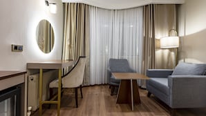 Een hotelkamer met een bureau, twee stoelen, een bank, een lamp, een spiegel en een raam met gordijnen.