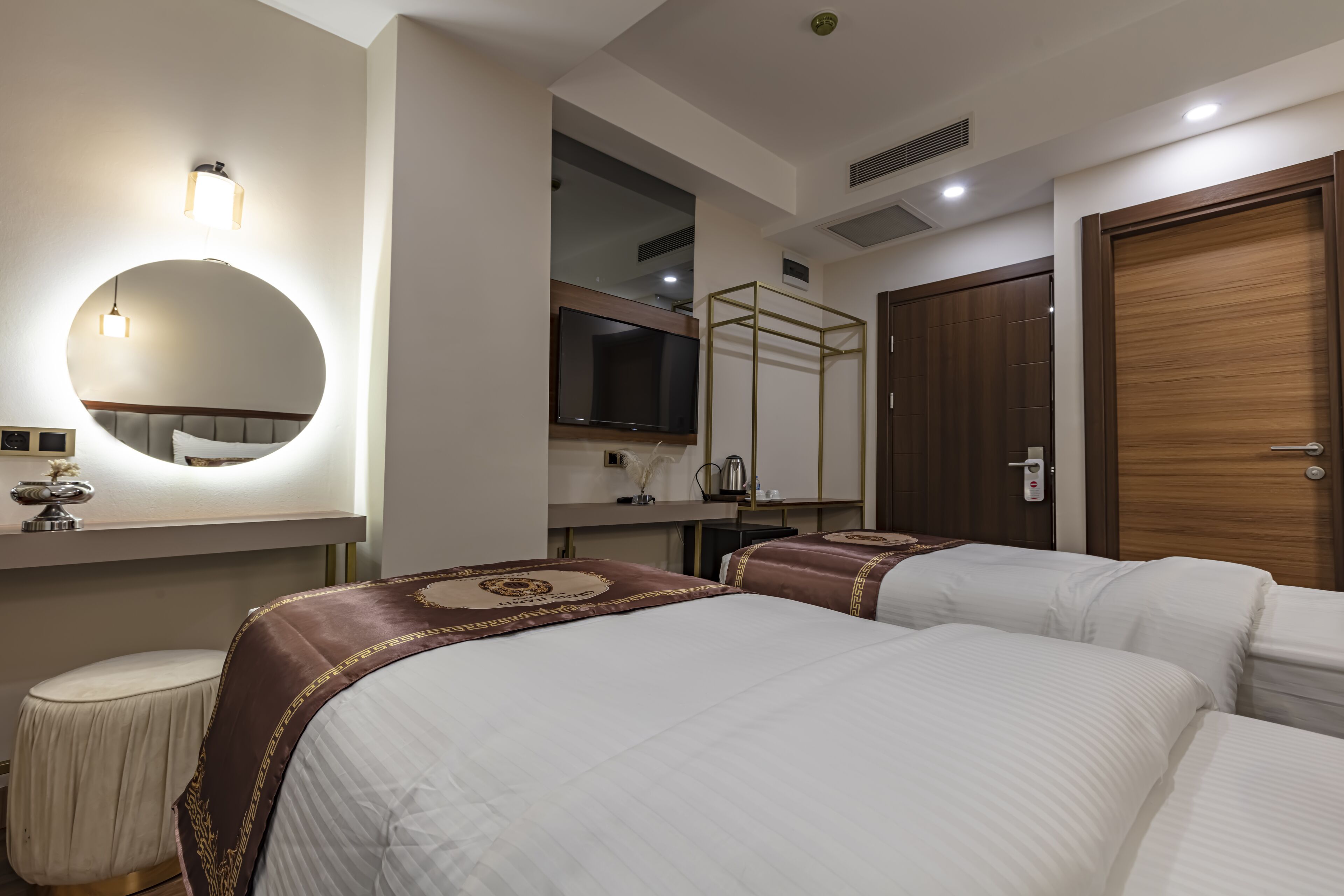 Habitación de hotel con dos camas, un espejo redondo, televisor, armario y una mesita de noche con lámpara.