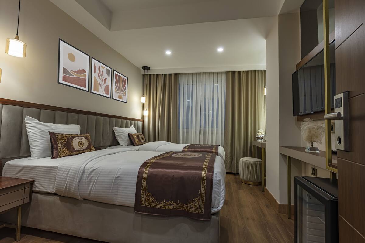 A hotel room with two beds, a bedside table, a lamp, a mirror, and a television.