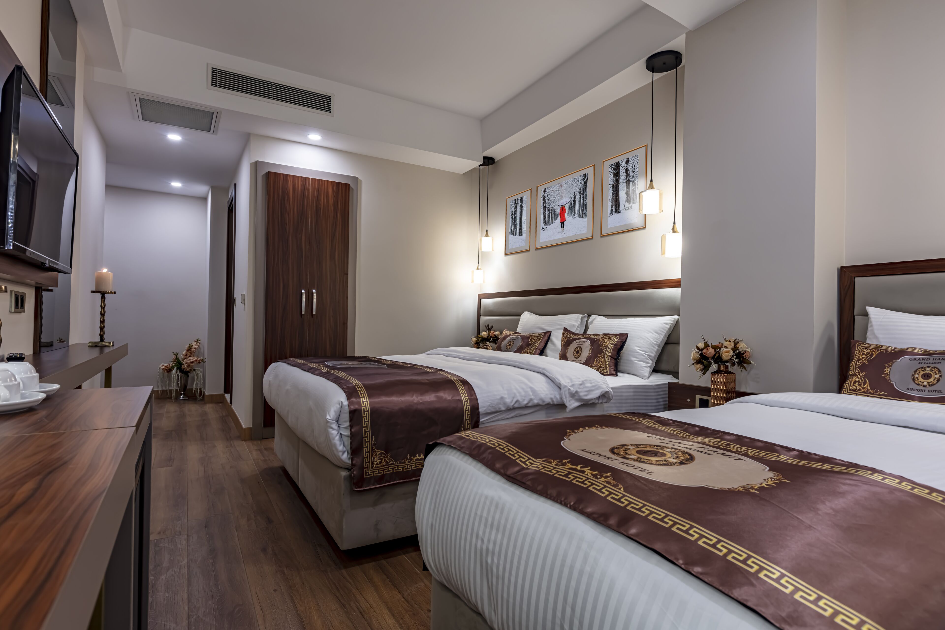 A hotel room with two beds, a TV, a desk with a lamp, and framed pictures on the wall.