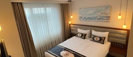 A hotel room with a large bed, a desk, a chair, a painting on the wall, and a window with curtains.