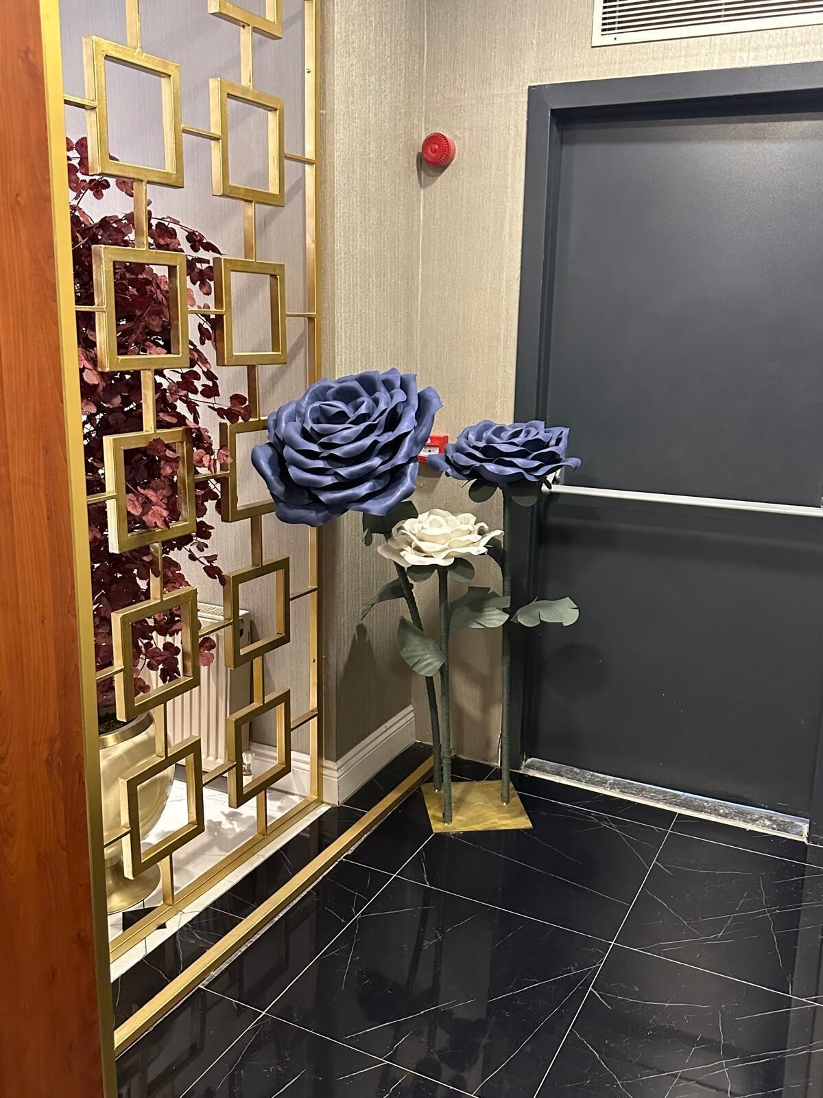 Rincón de una habitación con suelo de baldosas negras, pared decorativa con diseños geométricos y dos flores artificiales.