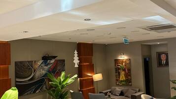 A modern hotel lobby with a seating area featuring sofas and armchairs, a large abstract painting, a potted plant, and a chandelier.