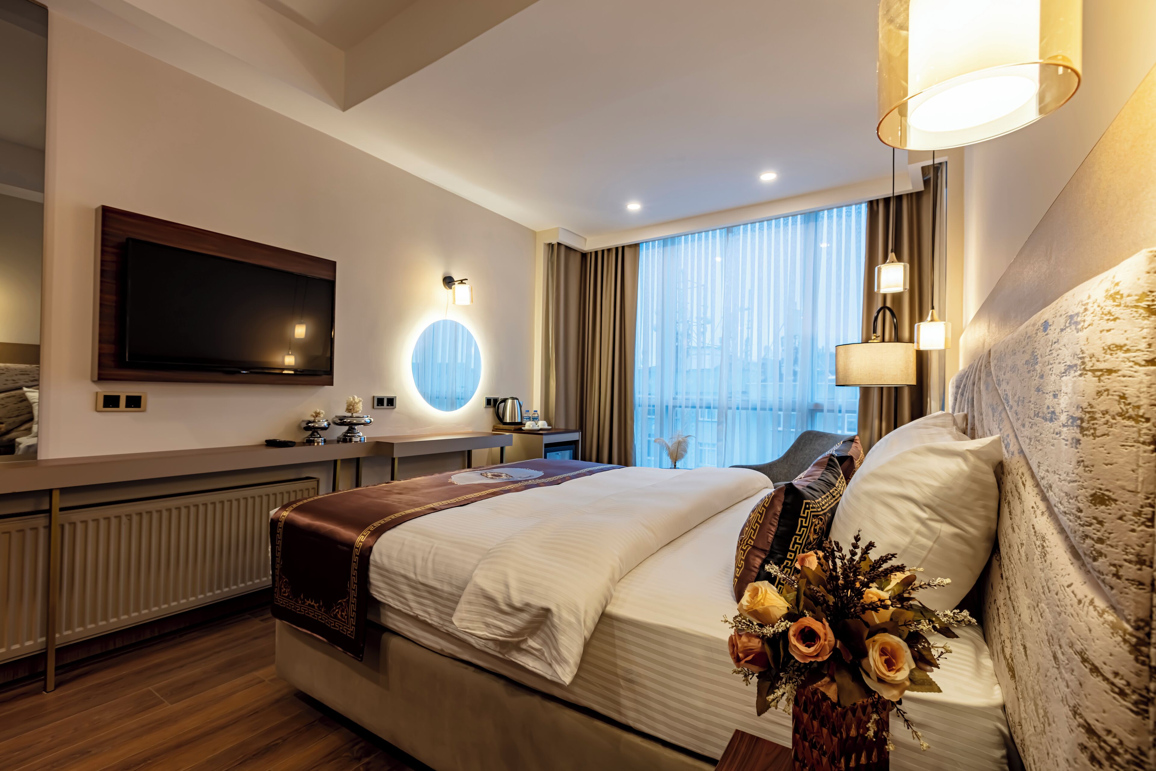 Habitación de hotel moderna con una cama grande, un televisor de pantalla plana, un escritorio con lámpara y vistas a la ciudad a través de amplias ventanas.