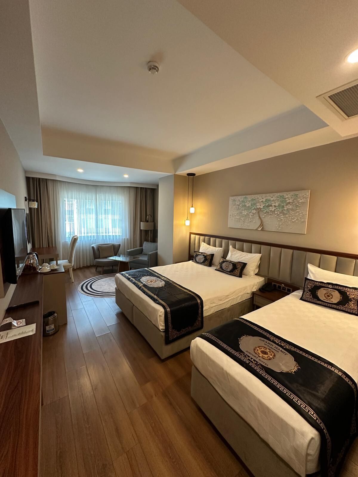 Habitación de hotel con dos camas, un escritorio, una silla y un ventanal grande con cortinas.