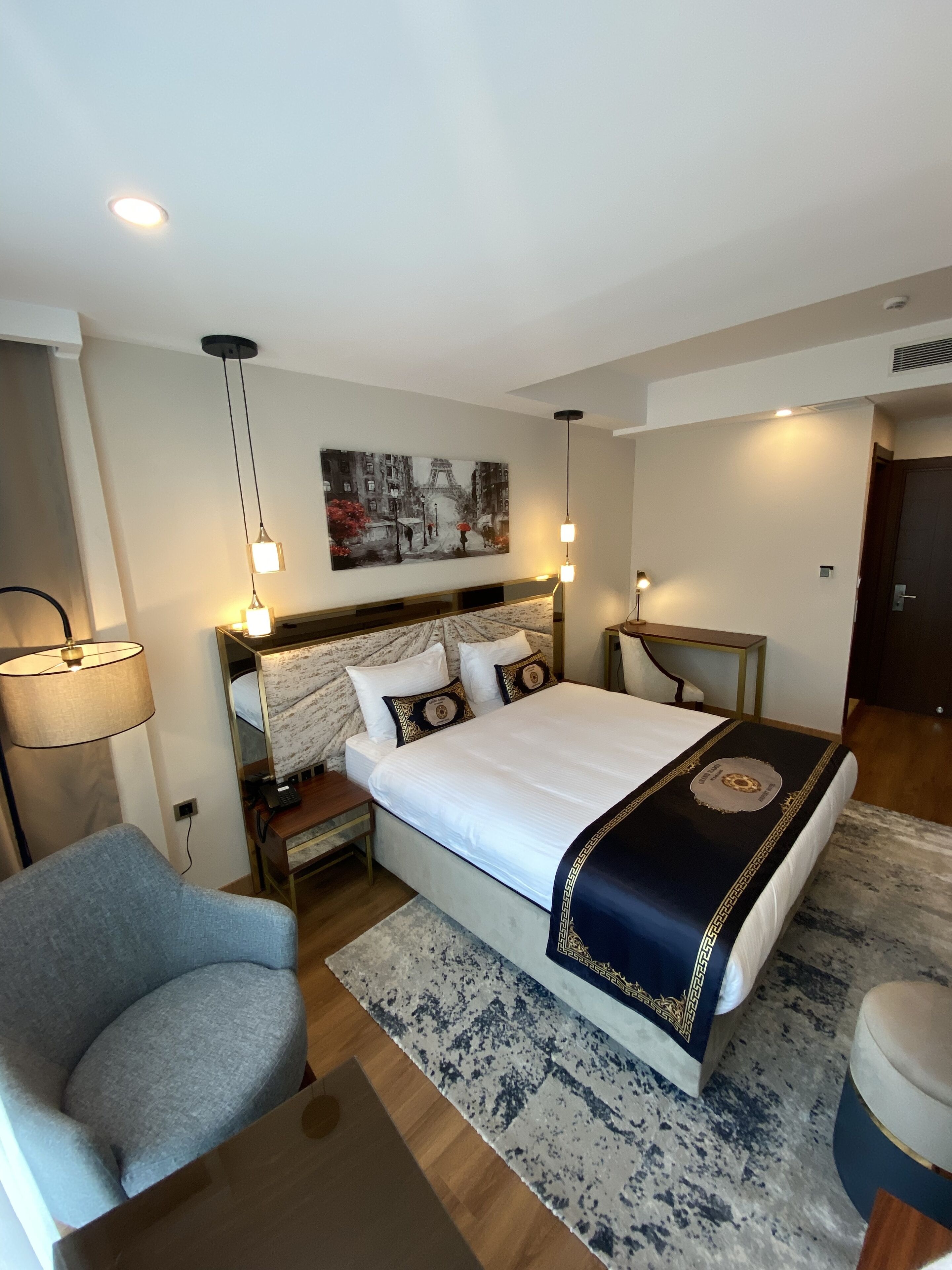 A modern hotel room with a large bed, a desk, a chair, and a painting of a cityscape.