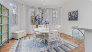 Un coin repas avec une table ronde, des chaises blanches, un tapis à motifs bleus et blancs, un banc blanc et une sculpture bleue.