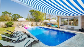 Un patio trasero con piscina, sillas de descanso, sombrillas y un área de terraza cubierta.