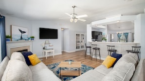 Una sala de estar moderna con sofá, mesa de centro y ventilador de techo. Hay una zona de cocina con taburetes de bar y un televisor montado en la pared.