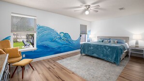Un dormitorio con cama, escritorio, silla, ventilador de techo y un mural en la pared.