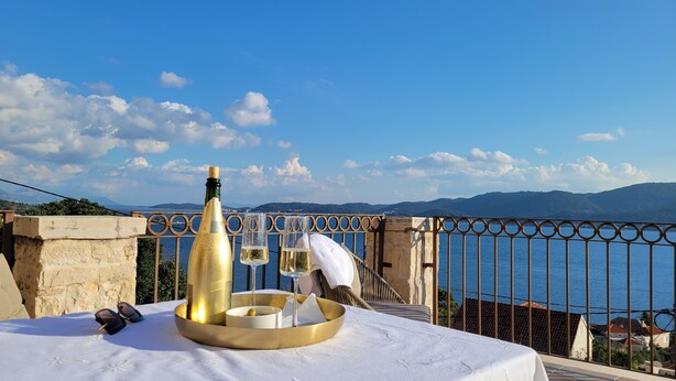 Un tavolo con una bottiglia, un bicchiere e una ciotola su un balcone affacciato su un corpo d'acqua e colline lontane.