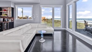 Ein großzügiger Wohnraum mit einem großen, weißen Ecksofa, einem Glastisch und Blick auf den Strand durch große Fenster.