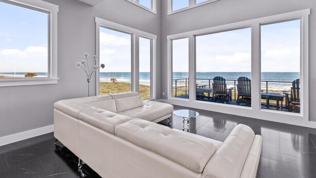 Ein modernes Wohnzimmer mit einem großen, weißen Ecksofa, einem runden Glastisch und Blick auf den Strand durch bodentiefe Fenster.