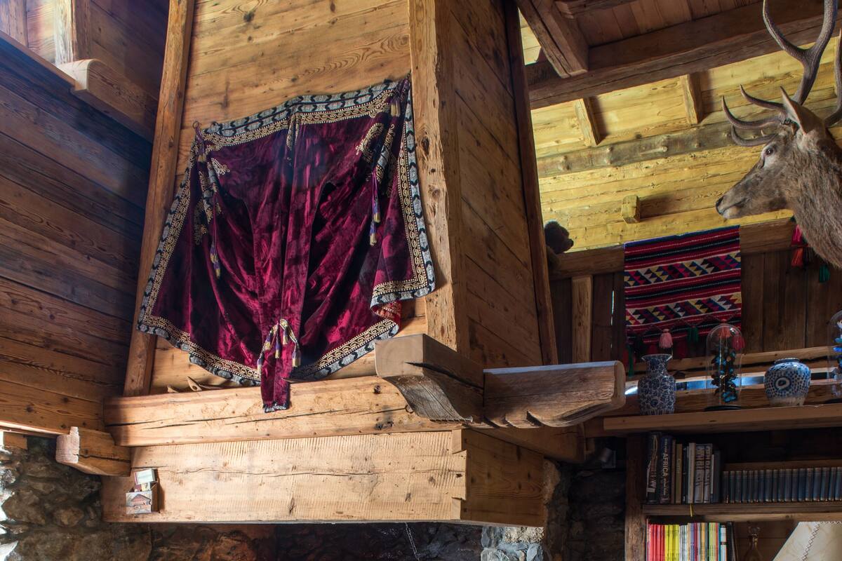 A cozy log cabin interior with a stone fireplace, a wooden ceiling, a bookshelf, and a deer head mounted on the wall.