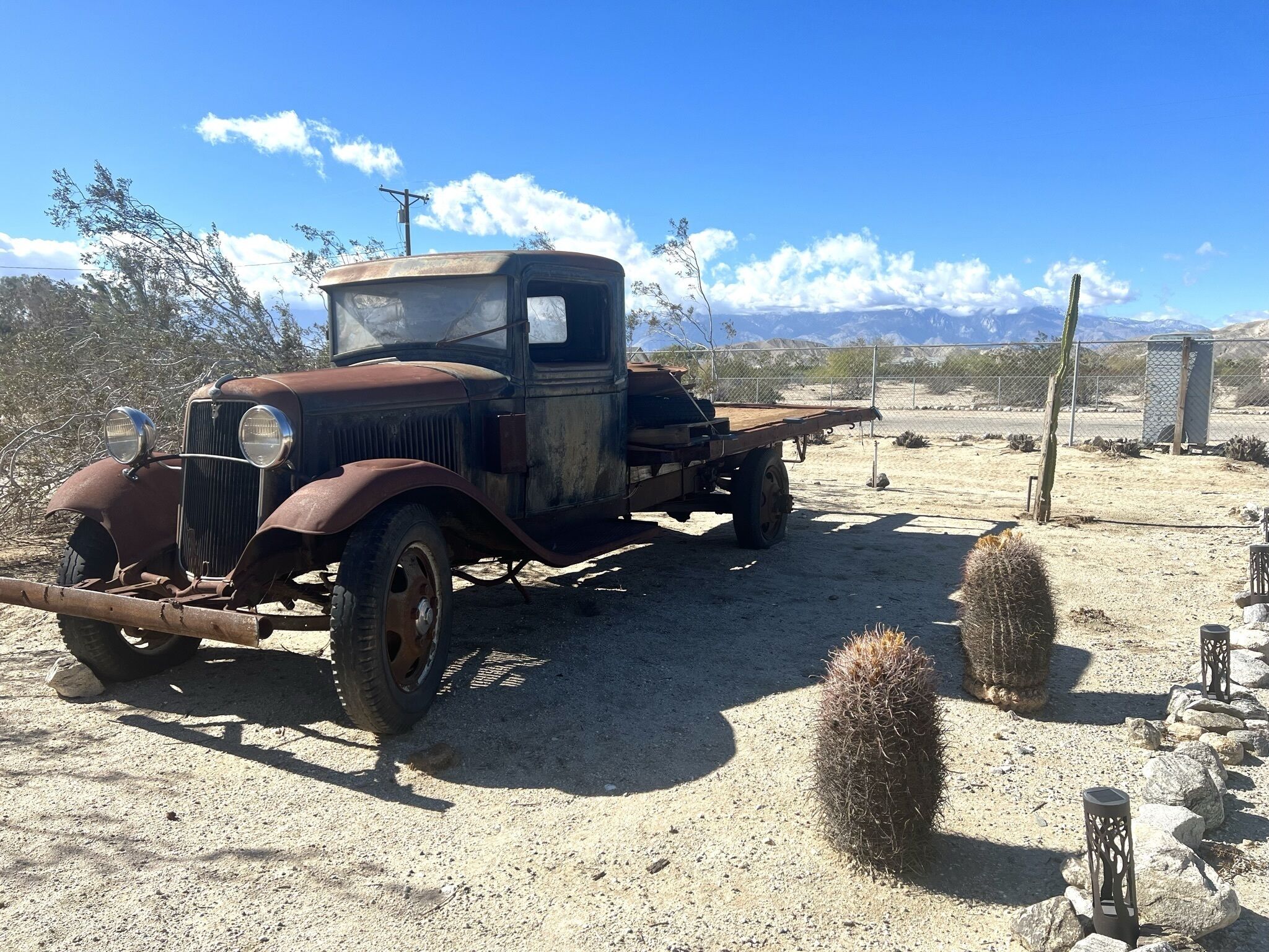 An old, rusty truck parked in a desert-like area with sparse vegetation and a clear sky.