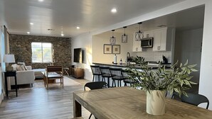 A modern living room with a stone wall, wooden furniture, and a dining area with a large table and bench. - Modern Ranch House~Great for Big Groups! Salt Water Pool + Spa  (Desert Hot Springs)