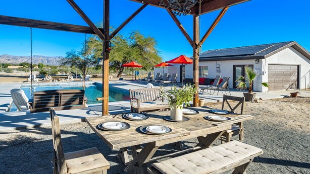 An outdoor pool area with a wooden pergola, lounge chairs, and a dining table set for a meal. - Modern Ranch House~Great for Big Groups! Salt Water Pool + Spa  (Desert Hot Springs)