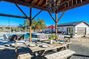 An outdoor pool area with a wooden pergola, lounge chairs, and a dining table set for a meal. - Modern Ranch House~Great for Big Groups! Salt Water Pool + Spa  (Desert Hot Springs)