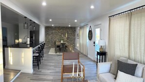 A modern living room with a sofa, a coffee table, and a dining area with a stone wall and a chandelier. - Modern Ranch House~Great for Big Groups! Salt Water Pool + Spa  (Desert Hot Springs)