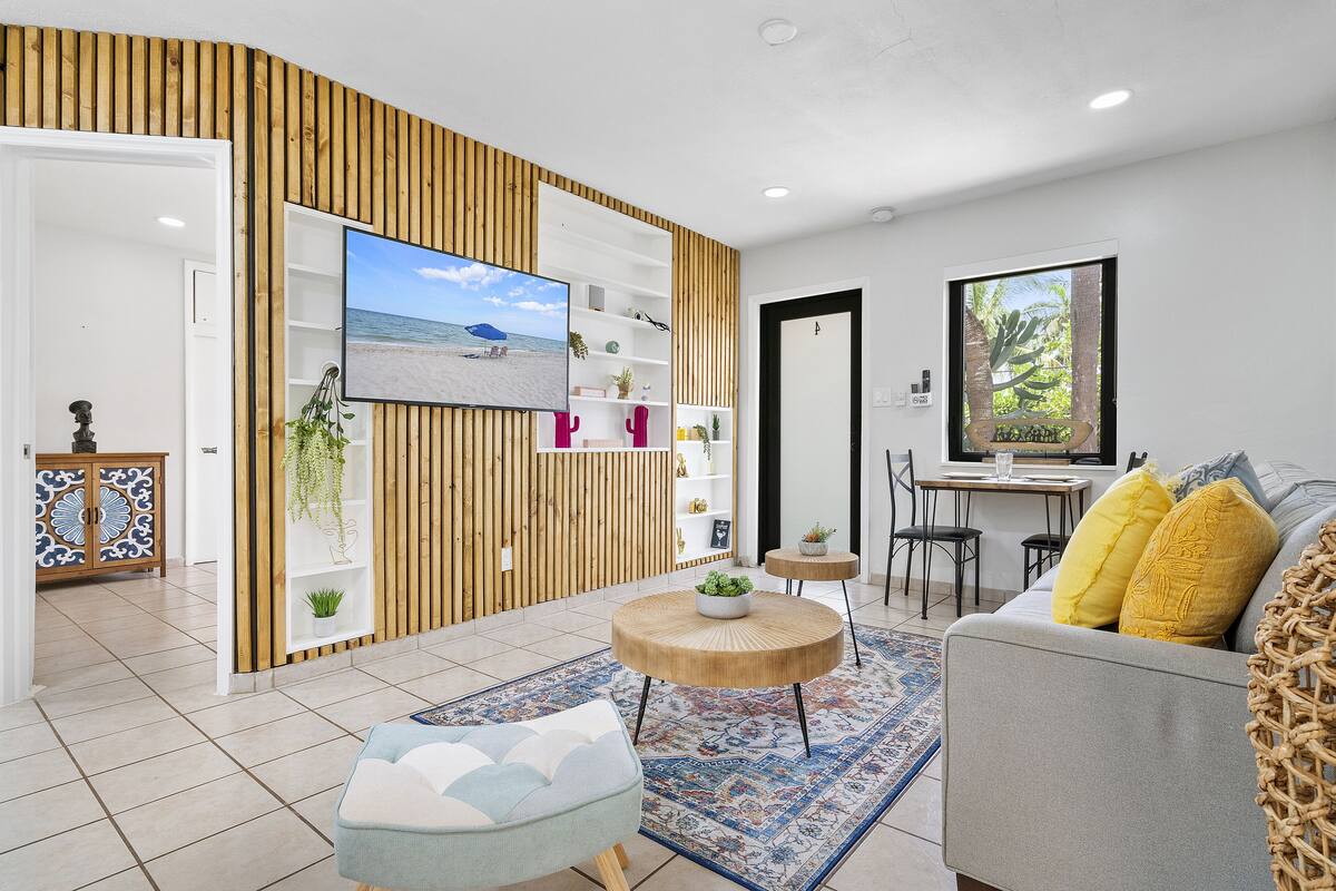 Una sala de estar moderna con un televisor de pantalla plana, un sofá con cojines amarillos, una mesa de centro redonda de madera y una alfombra estampada.