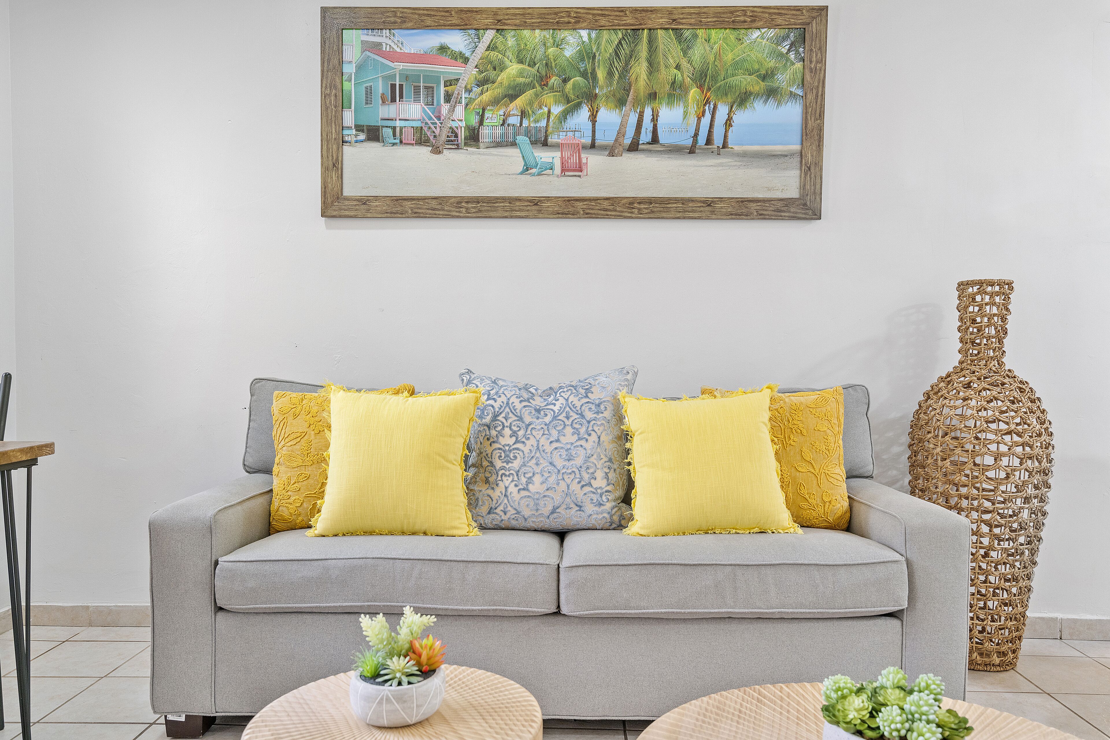 Una sala de estar con un sofá gris, cojines estampados en amarillo y azul, un florero de mimbre y un cuadro de una playa.