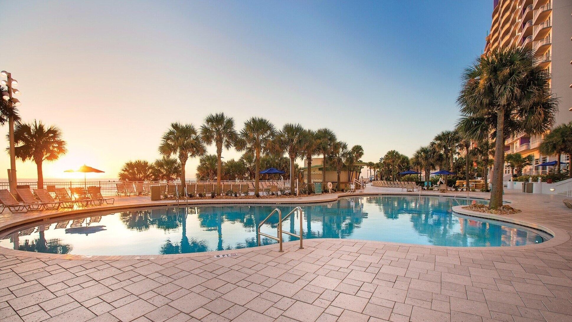 A swimming pool with lounge chairs and palm trees, surrounded by a hotel building.