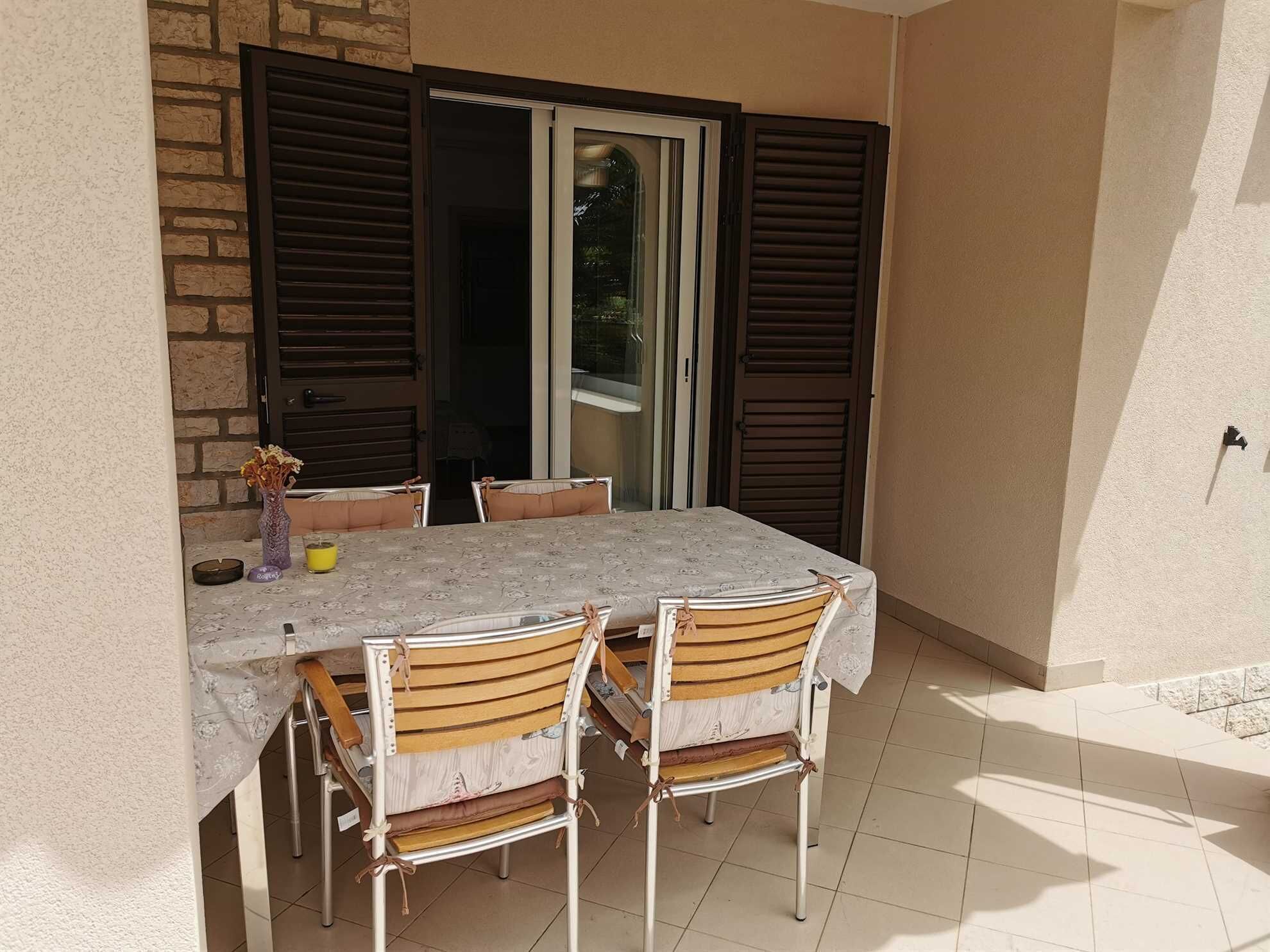 Eine Terrasse mit Tisch und Stühlen, einer Blumenvase und einem Fenster mit Rollläden.