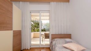Ein Schlafzimmer mit hölzernem Kopfteil, einem Bett mit gemusterten Betttextilien, einem Fenster mit weißen Vorhängen und Blick auf einen Balkon mit Stuhl und Tisch.