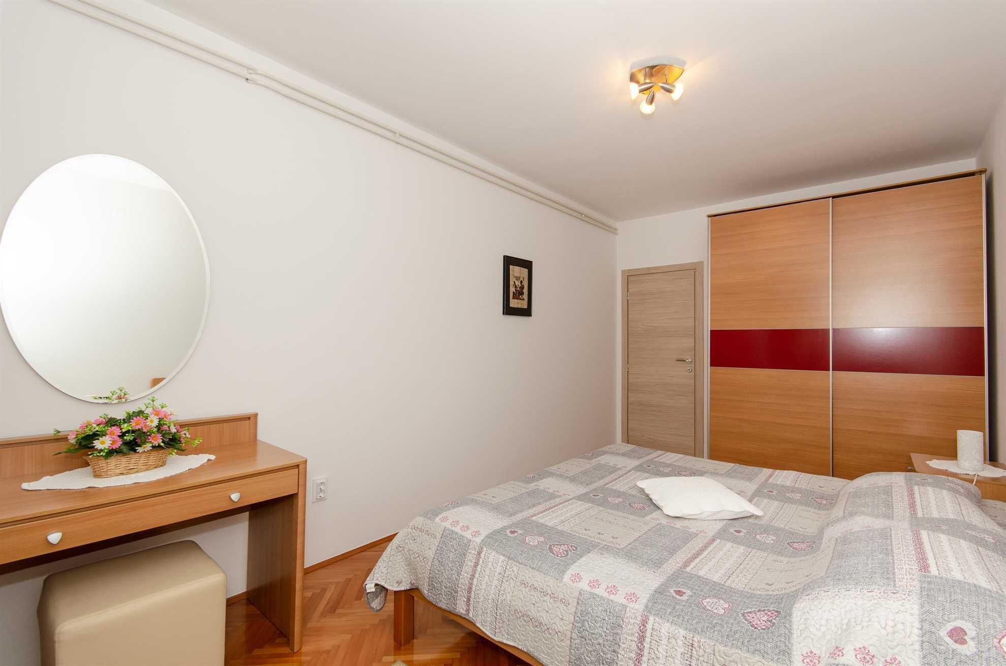 A bedroom with a wooden desk, a large mirror, a bed with a patterned quilt, and a wardrobe with a red stripe.