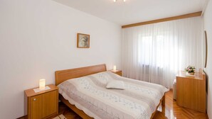 Ein Schlafzimmer mit einem hölzernen Bett, Nachttischen, einer Nachttischlampe, einem gerahmten Bild und einem Fenster mit durchsichtigen Vorhängen.