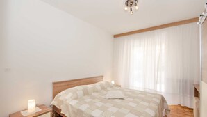 A bedroom with a wooden headboard, a bedside table with a lamp, and a large window with sheer curtains.