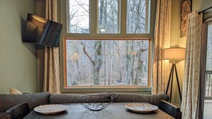 A dining area with a table, chairs, a TV mounted on the wall, a window with a view of trees, and a lamp.