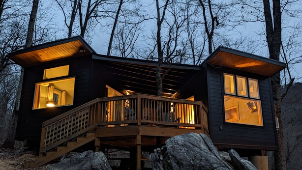A cabin with a wooden deck, large windows, and a staircase leading to the entrance.