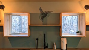 A bathroom with two windows, a sink, a mirror, a towel dispenser, and a small plant.